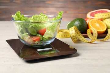 Scales with salad, products and measuring tape on white wooden table, closeup