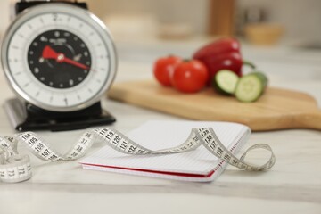 Kitchen scales, measuring tape, notebook and vegetables on light marble table indoors, closeup