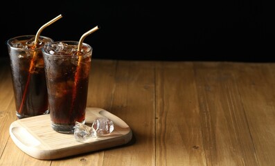 Refreshing cola with ice cubes and straws in glasses on wooden table against black background. Space for text