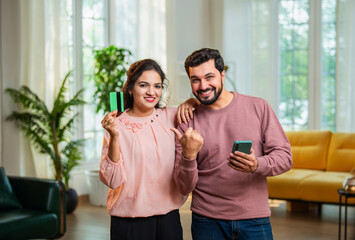 Indian man and woman cheerfully showing credit card and phone in living room after reward