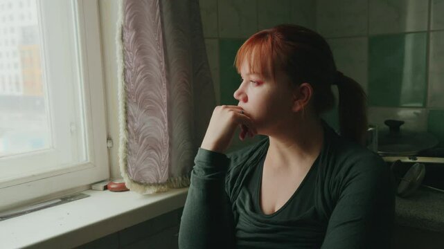 A young woman with red hair sits quietly by a window, lost in thought while observing the world outside