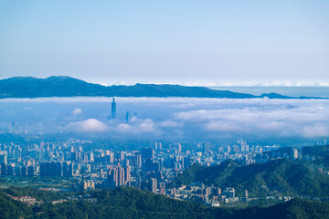 Fototapeta premium A serene spring morning view of Taipei from Lion Head Mountain. White clouds drift over the city skyline, featuring prominent high-rise buildings. 