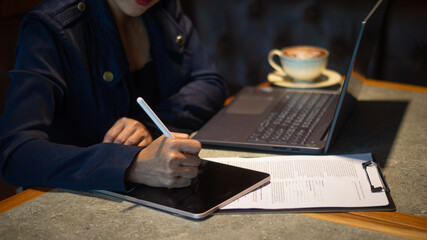 A businesswoman is intently writing something on a tablet placed on a checklist, using a white touchscreen pen, and working on a laptop in a cozy corner of a cafe.