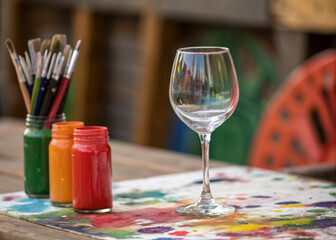 Colorful paintbrushes in a jar stand beside a wine glass filled with a light liquid on a table covered in artistic paint splatters