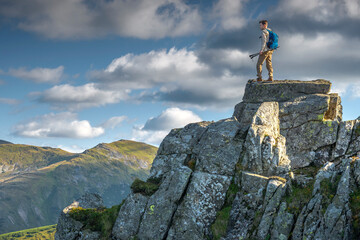 Male hiker standing on top of a rocky cliff in the mountains with trekking poles, looking at the...