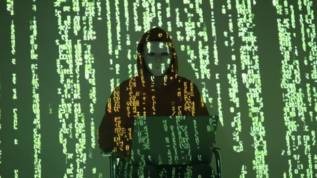 Zamaskowany haker w ciemnym kapturze w otoczeniu zielonego kodu matrix pracuje na laptopie &ndash; cyberprzestępczość, anonimowość w sieci, atak hakerski, cyberbezpieczeństwo i kodowanie

