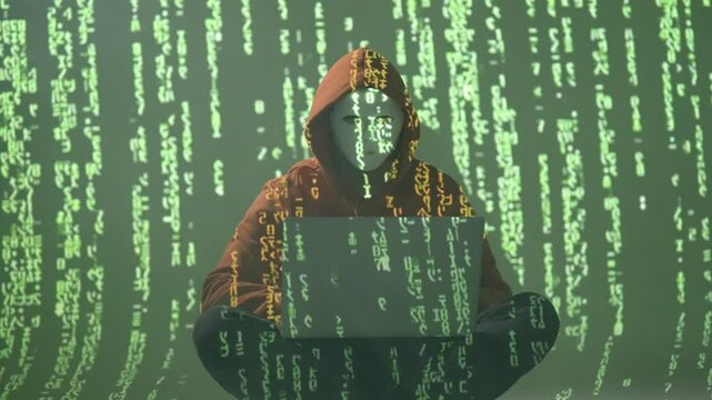 Anonimowy haker w masce i kapturze z laptopem na tle zielonego kodu matrix &ndash; cyberprzestępczość, programowanie, zagrożenia sieciowe i cyfrowa inwigilacja w mrocznym środowisku