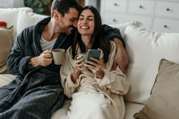 Young couple wearing bathrobes enjoying a relaxing morning on the sofa, drinking coffee and using a smartphone