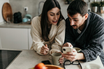 Young couple wearing bathrobes looking with surprise at a smartphone in a modern kitchen in the morning