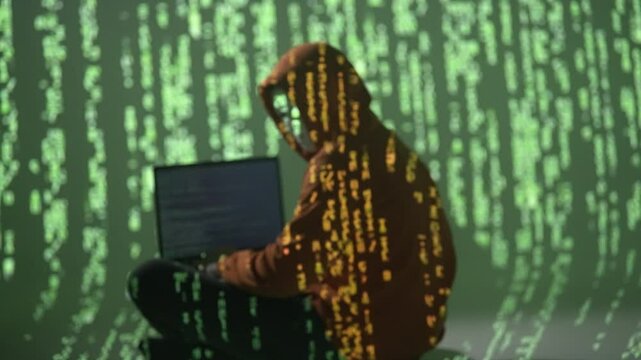 Anonimowy haker w masce i kapturze przy komputerze, z zielonym kodem matrix na tle, symbolizujący cyberatak, nielegalne działania online i zagrożenia bezpieczeństwa cyfrowego