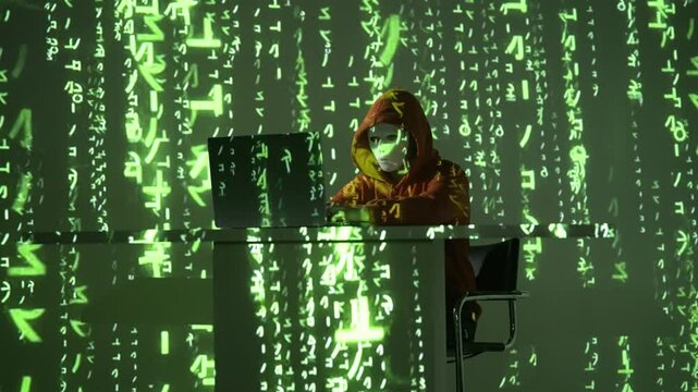 Cyberprzestępca w masce i kapturze przy laptopie otoczony zielonym kodem matrix, siedzący w ciemnym pokoju, symbolizuje hakera w akcji, cyberatak i nowoczesne zagrożenia cyfrowe