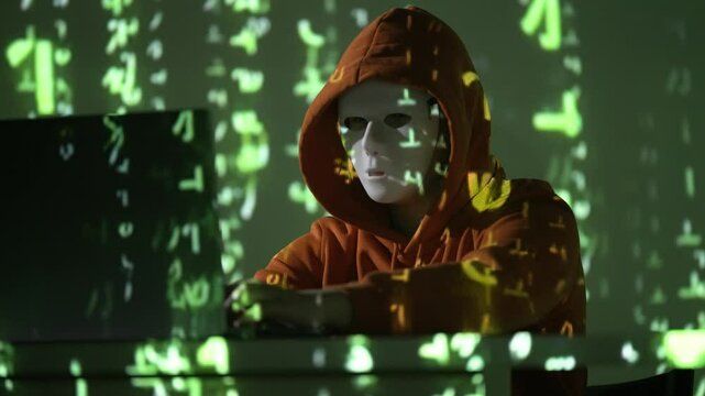 Zamaskowany cyberprzestępca w kapturze pracuje przy laptopie, otoczony zielonym cyfrowym kodem matrix, symbolizującym hakowanie, tajne operacje i zagrożenia bezpieczeństwa w sieci