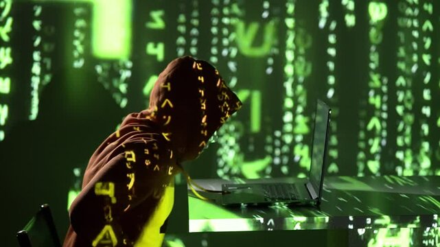 Haker w kapturze pochyla się nad laptopem w ciemnym pokoju z projekcją zielonego kodu matrix, przygotowując się do cyberataku w cyfrowym świecie pełnym zagrożeń i anonimowości