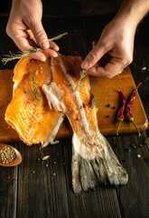 Fresh salmon fillet rests on a wooden cutting board as hands sprinkle herbs and spices. Red chilies...