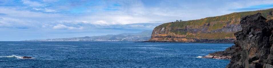 Fototapeta premium Blick von Água de Pau in Richtung Ponta Delgada (Azoren). Im Vordergrund massive Lavafelsen und blaues Meer, dazwischen die Bucht Baixa d'Areia Beach. Im Hintergrund Ponta Delgada unter blauem Himmel.