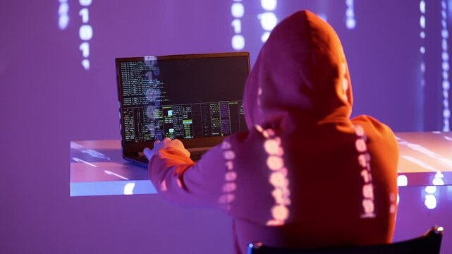 Postać w kapturze analizująca kod systemowy na laptopie z czerwonymi projekcjami binarnymi w tle