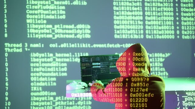 Osoba w kapturze przed laptopem z kodem na ekranie, otoczona zielonymi danymi systemowymi &ndash; haker, system, cyberatak, analiza, bezpieczeństwo