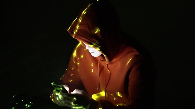 Anonimowa postać w masce i kapturze w ciemności, otoczona ż&oacute;łtym kodem, wpatrzona w telefon &ndash; haker, dane, cyberświat, zagrożenie