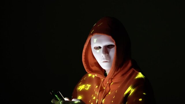 Postać w masce i pomarańczowej bluzie skupiona na telefonie, oświetlona ż&oacute;łtym kodem &ndash; haker, anonimowość, cyberświat, dane