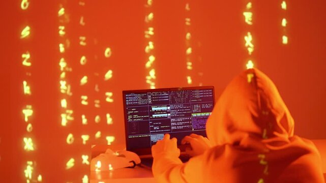 Haker w kapturze pracujący przy laptopie z kodem, otoczony projekcją binarną &ndash; dane, cyberatak, kodowanie, cyberbezpieczeństwo