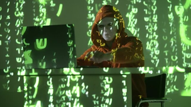 Osoba w masce i kapturze przy laptopie w otoczeniu zielonego kodu &ndash; haker, cyberbezpieczeństwo, dane, anonimowość, cyberatak