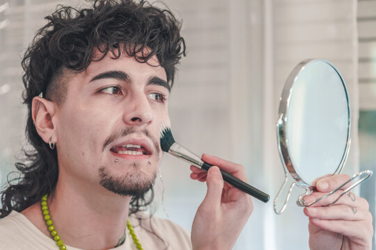 Young man applying makeup with brush in bathroom mirror: embracing diversity and inclusion