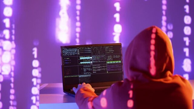 Osoba w kapturze pracująca przy laptopie, otoczona projekcją kodu binarnego &ndash; cyberatak, kodowanie, dane, haker, systemy komputerowe