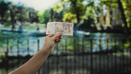 Man holding saudi riyal banknote in outdoor park setting with blurred background depicting nature and leisure scene, emphasizing currency and location.