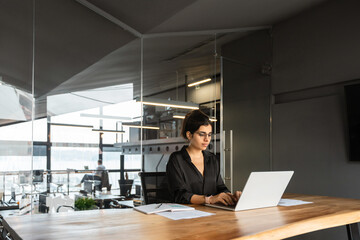 Young middle eastern or indian businesswoman in formal clothes and eyeglasses sitting at desk workplace working on computer. Latin business entrepreneur woman it specialist typing, browsing in office