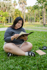 chubby asian woman sits on green grass in park, reading book with focus and calmness, surrounded by...