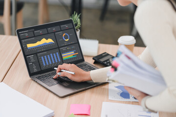 Businesswoman analyzing data report on laptop dashboard with charts and graphs, holding documents in modern office.