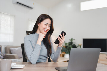 Obraz premium Asian businesswoman smiling while using smartphone and working on laptop in modern office setting
