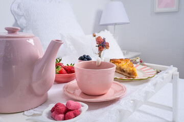 Detail of a delicious breakfast on a bed in white. Natural light enters through the window. There is a teapot, a tray, fruit , a sprig of flowers and an apple pie. Care and love