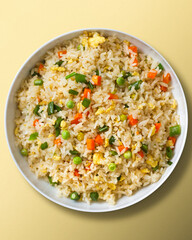 Top view of a bowl with fried rice with egg and vegetables, onion, bell pepper, carrot, green peas. Vegetarian food served in a white bowl on a beige