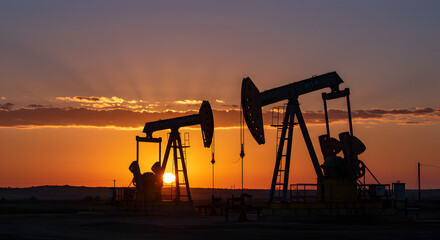 Naklejka premium Silhouetted Oil Pumps Against Setting Sun in Rural Landscape with Warm Golden Sky and Industrial Machinery