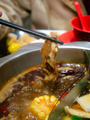human hand using chopsticks hold meat over Sichuan mala hotpot shabu on restaurant table.