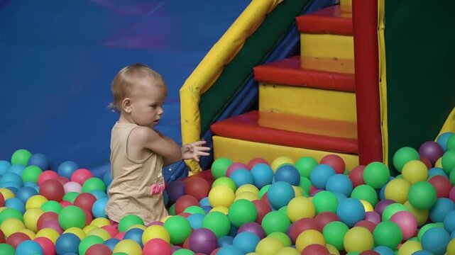 little girl beautiful T-shirt shorts sits pool balls children's playground and takes turns taking colorful balls throwing them far, clearing her way steps, warm summer day, childhood, enjoyment, joy
