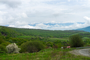 Part of asphalt mountain road, road signs. Fields with green grass and bushes on the hills. Blue sky with thick clouds in the background