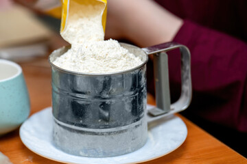 Pouring wheat flour into a metal cup sieve for precise sifting, capturing a key baking preparation step in rustic style