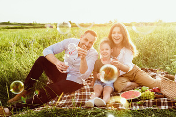 Picnic Family Fun. Adorable girl with her mom and dad blowing soap bubbles in coutryside on summer day