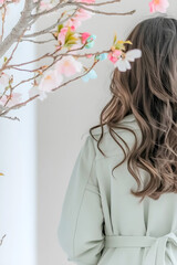 Naklejka premium Woman with long wavy hair in a light coat standing near blooming cherry blossoms in soft pastel light