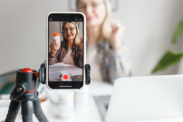 Selective focus on phone on tripod recording video of senior qualified nutritionist female blogger of 60s holding bottle of food supplement, giving advice and tips on how boost health, immune system
