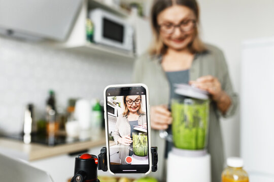 Selective focus on smartphone screen recording video recipe tutorial of popular snior female vlogger or blogger, influencer, standing at kitchen with blender, making detox smoothie to stay healthy - Powered by Adobe