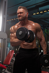 Naklejka premium athletic man engages in powerful strength training session showcasing his muscular physique while lifting heavy dumbbells inside modern gym. atmosphere is energetic and intense.