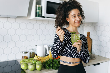 Cute funny african american girl of generation z eating smoothie with spoon in kitchen, standing next to counter, looking at camera like saying want some offering to try healthy delicious snack