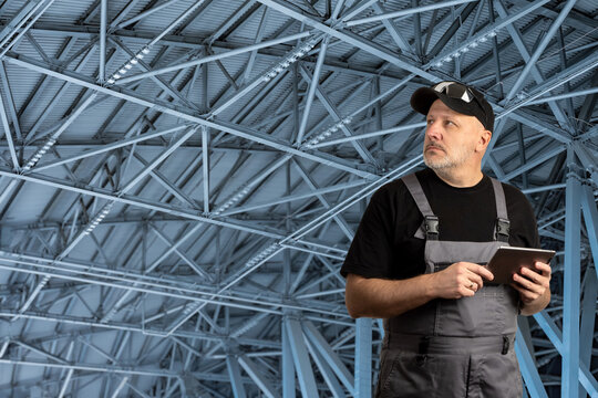 Construction specialist inspects a metal truss roof structure with a digital tablet, focusing on structural integrity and building engineering tasks.
