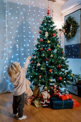 Little girl stands with a teddy bear near a decorated Christmas tree and points to the top. Back view