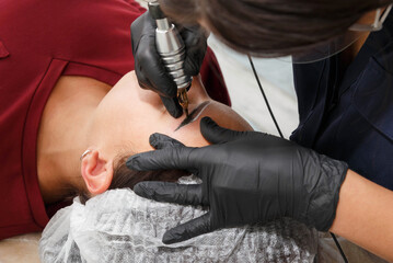 The master applies eyebrow makeup paint to the client with a tattoo machine