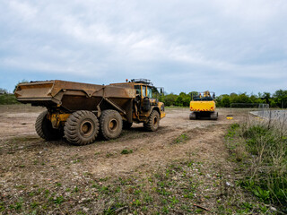Obraz premium Heavy-duty construction vehicles, including a dump truck and excavator, parked on a dirt worksite surrounded by nature under a cloudy sky, ready for earthmoving tasks
