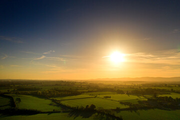 View of lush green farmland at golden hour, with long shadows cast by hedgerows and trees, glowing fields, and distant hills softly lit by the warm setting sun under a clear sky.